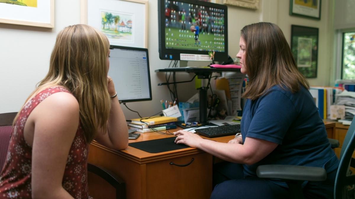 Professor talking to student in her office while typing on computer